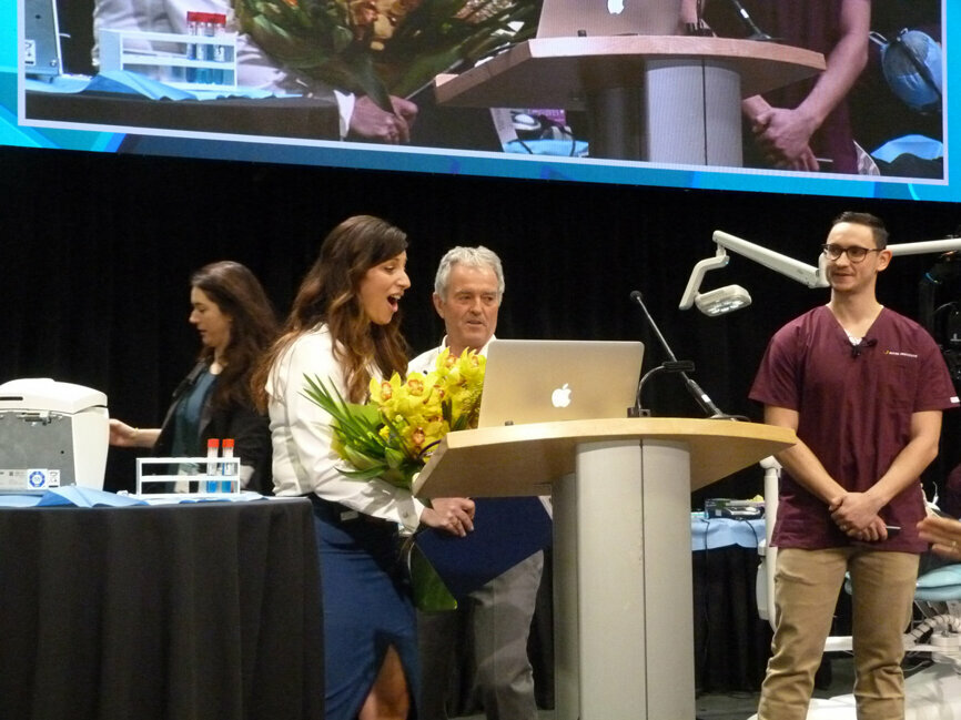 Penny Hatzimanolakis, RDH, is surprised Thursday morning with a bouquet of flowers in honor the 10th season of the Live Dentistry Stage at PDC — and Hatzimanolakis’s 10th season of hosting. Presenting the flowers on behalf of the PDC organizing committee is Bruce Ward, DDS, chair of the committee. Also on stage is Dmitriy Ivanov, DDS, about to present a live demonstration with Ho-Young Chung, DDS. Hatzimanolakis has been on the PDC organizing committee since 2007, and among here suggestions for improving the Live Dentistry Stage this year was ‘adding some flowers’ out in front. (Photo: Robert Selleck/Dental Tribune)